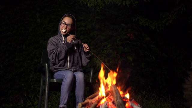 一个戴兜帽的年轻女人在火上烤棉花糖。晚上篝火。视频素材