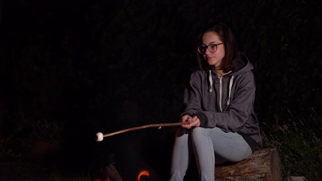 一个戴兜帽的年轻女人在火上烤棉花糖。晚上篝火。视频素材