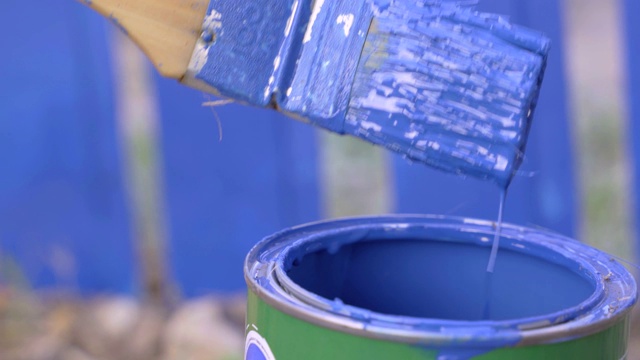 勤杂工将刷子蘸入蓝色染料的金属罐中，小心地摇掉多余的水滴，在建筑工地的栅栏或墙壁上或村庄的土地上，靠近视频下载