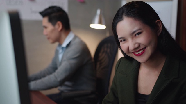 迷人的亞洲女商人在電腦上工作，微笑著看著相機，積極，滿意，愉快的表情享受在現代辦公室的成功擺姿勢。視頻素材