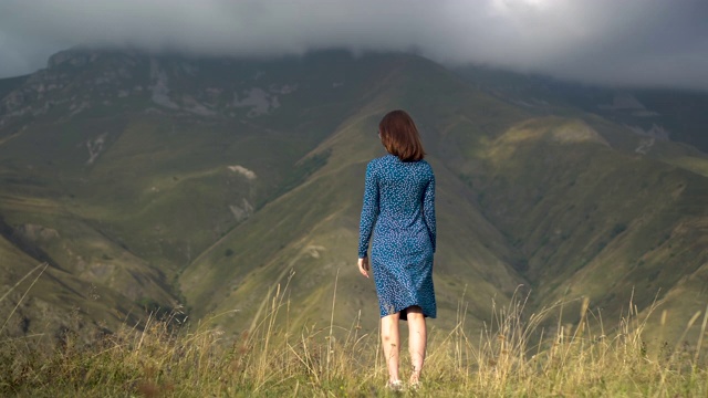一位身穿蓝色连衣裙的年轻女子站在那里，高举双手望着群山。视频下载