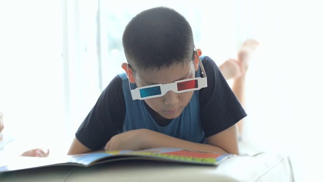 一个亚洲男孩躺在床上，戴着3d眼镜在家靠窗看书。视频下载