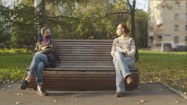 两个女人戴着一次性保护面罩或头巾，坐在长椅上保持社交距离视频下载