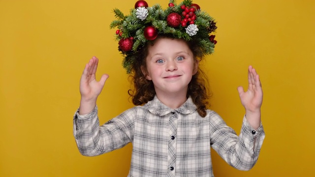惊喜可爱的小女孩用手摆出大尺寸的姿势，印象深刻的孩子展示出宽的东西，戴着手工制作的花环，孤立在黄色的工作室背景上。圣诞快乐的概念视频素材
