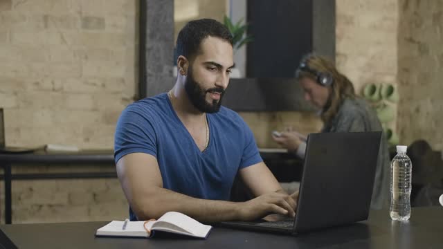 一幅自信的中东男学生在笔记本电脑键盘上打字的照片，背景是模糊的朋友在做作业。英俊的年轻人在网上学习。电影4k ProRes总部。视频下载