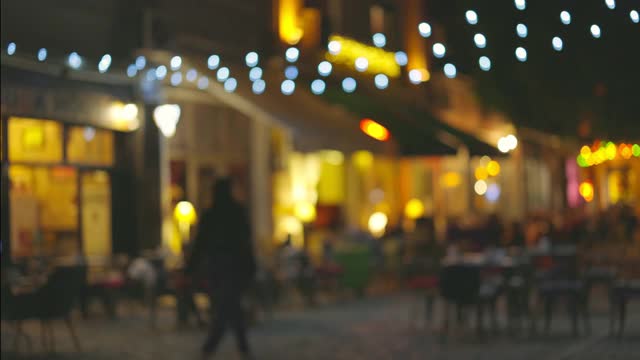慢镜头拍摄的灯火辉煌的街道与咖啡馆和夏日露台。人们沿着老城的步行街走，选择一个地方吃饭。浪漫之夜的温馨餐厅。模糊4 k拍摄视频素材