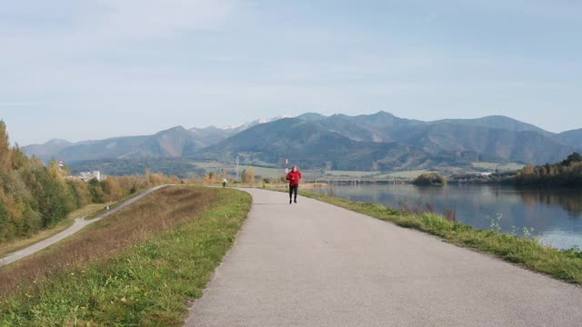 身穿红色长袖衬衫的男子在以山为背景的柏油路上奔跑的慢动作视频。运动的人的活动和健康的生活方式的概念镜头。视频素材