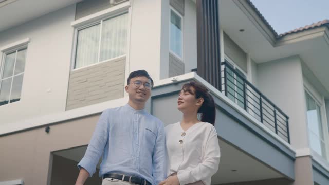 这是一对亚洲年轻夫妇站在他们的新房子前，拥抱在一起，手里拿着房子钥匙，看起来很高兴开始新的生活。家庭、年龄、住宅、房地产和人的概念。视频购买