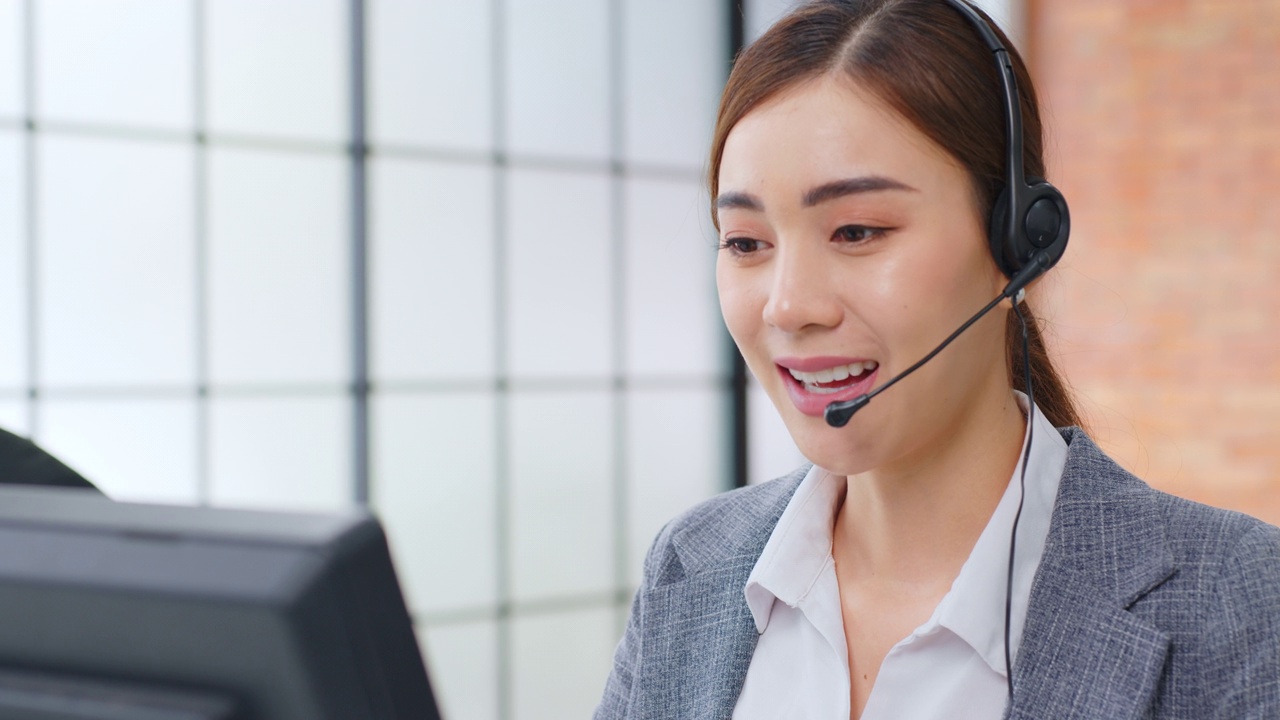 亚洲女性呼叫中心，客户服务，电话销售在正式的西装戴耳机或耳机与客户在现代办公室谈话视频购买