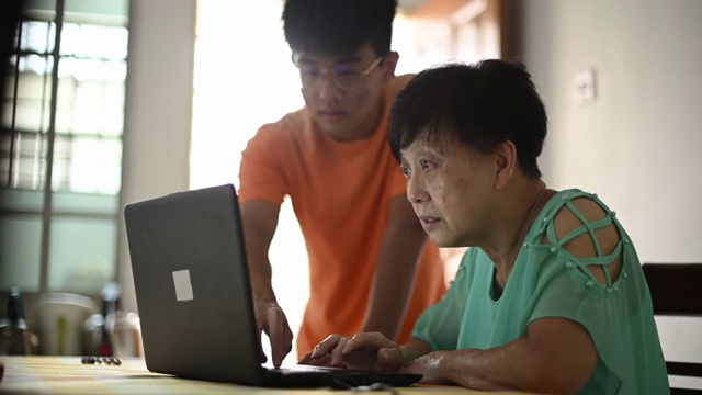 在餐厅里，一位年长的亚洲华人女士向她的孙子学习如何使用笔记本电脑社交媒体视频下载