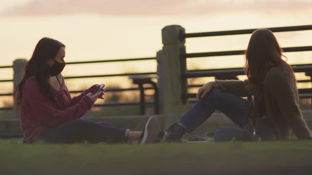 一名年轻女子给她的朋友拍照，他们坐在户外的草地上，戴着防护口罩视频素材