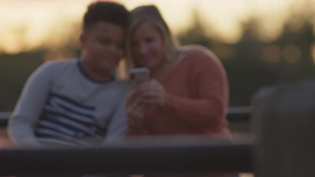 慈母和儿子坐在河边的户外一起看智能手机屏幕视频素材