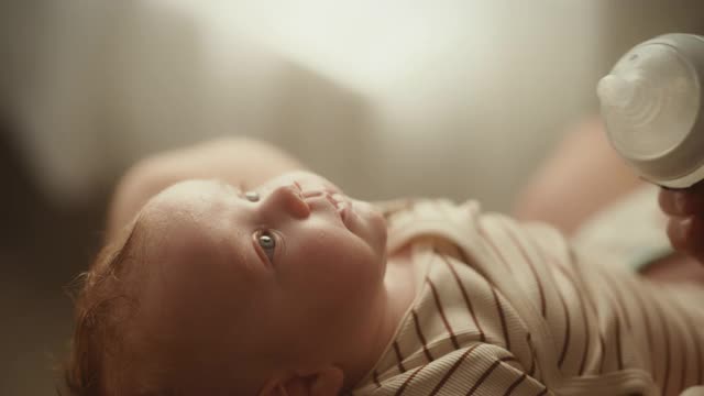 真实的近距离镜头，一个新生儿喝牛奶从小婴儿瓶，而躺在儿童床上。家庭中的白人新生儿的肖像。儿童与父母的概念。视频购买