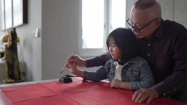 亚洲华人祖父和孙女在中国新年庆祝期间练习书法，家里装饰一张红色的纸视频下载