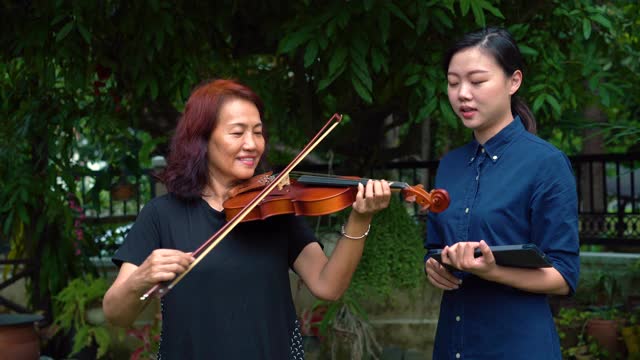 年轻的亚洲妇女教她的母亲如何在室外拉小提琴视频素材