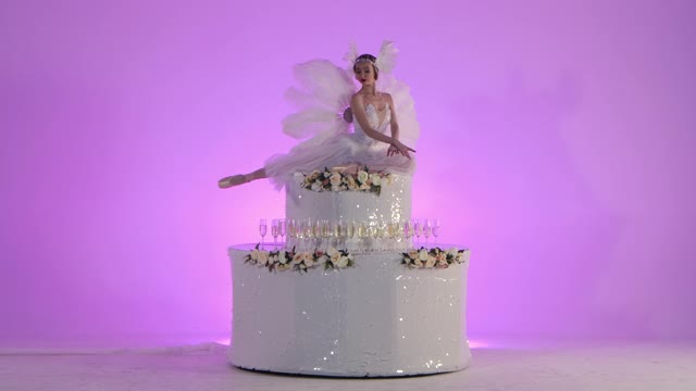 一个精致的女性芭蕾舞演员打扮成一只白天鹅，在一个装饰着鲜花的蛋糕上摆姿势。在演播室的粉色背景上，金发女郎优雅地慢动作。节日戏剧舞蹈表演视频下载