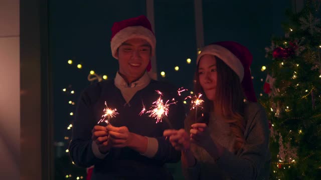 一对亚洲夫妇站在一个有圣诞树的房间里，在一个派对上用烟花。男人和女人在圣诞节和新年互相庆祝。视频下载