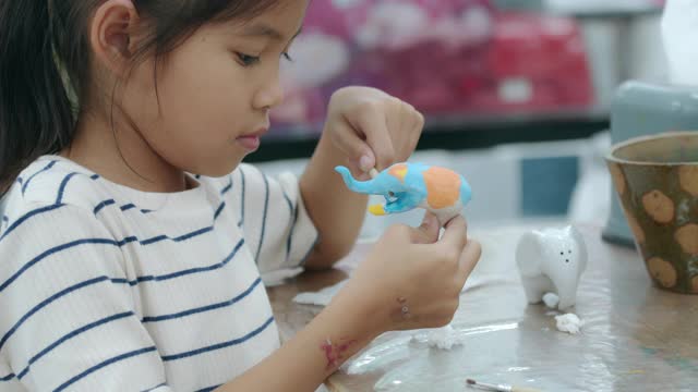 亚洲小女孩的慢镜头在小陶瓷象上兴致勃勃地画着油画。孩子们在学校的艺术和手工艺创意活动班。视频下载
