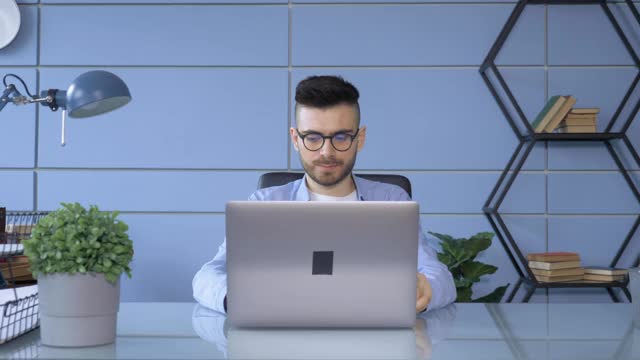 年轻男子在时尚眼镜看眼睛监视器，上网。在他的家庭办公室里用笔记本电脑工作的时尚男性视频素材