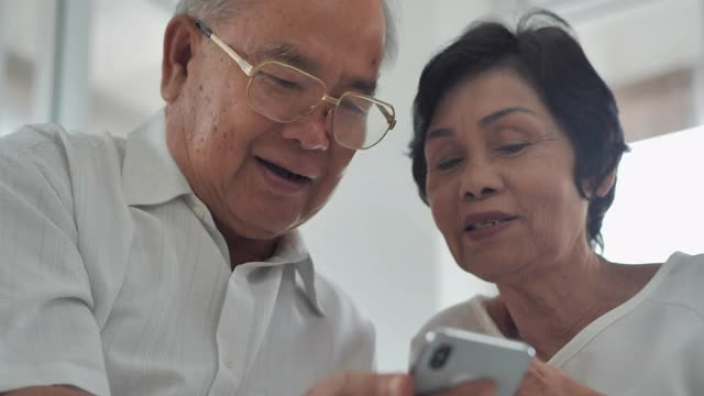 一对60-70岁的亚洲夫妇坐在养老院客厅的沙发上，一边聊天一边用智能手机看在线内容。高级技术的概念。视频下载