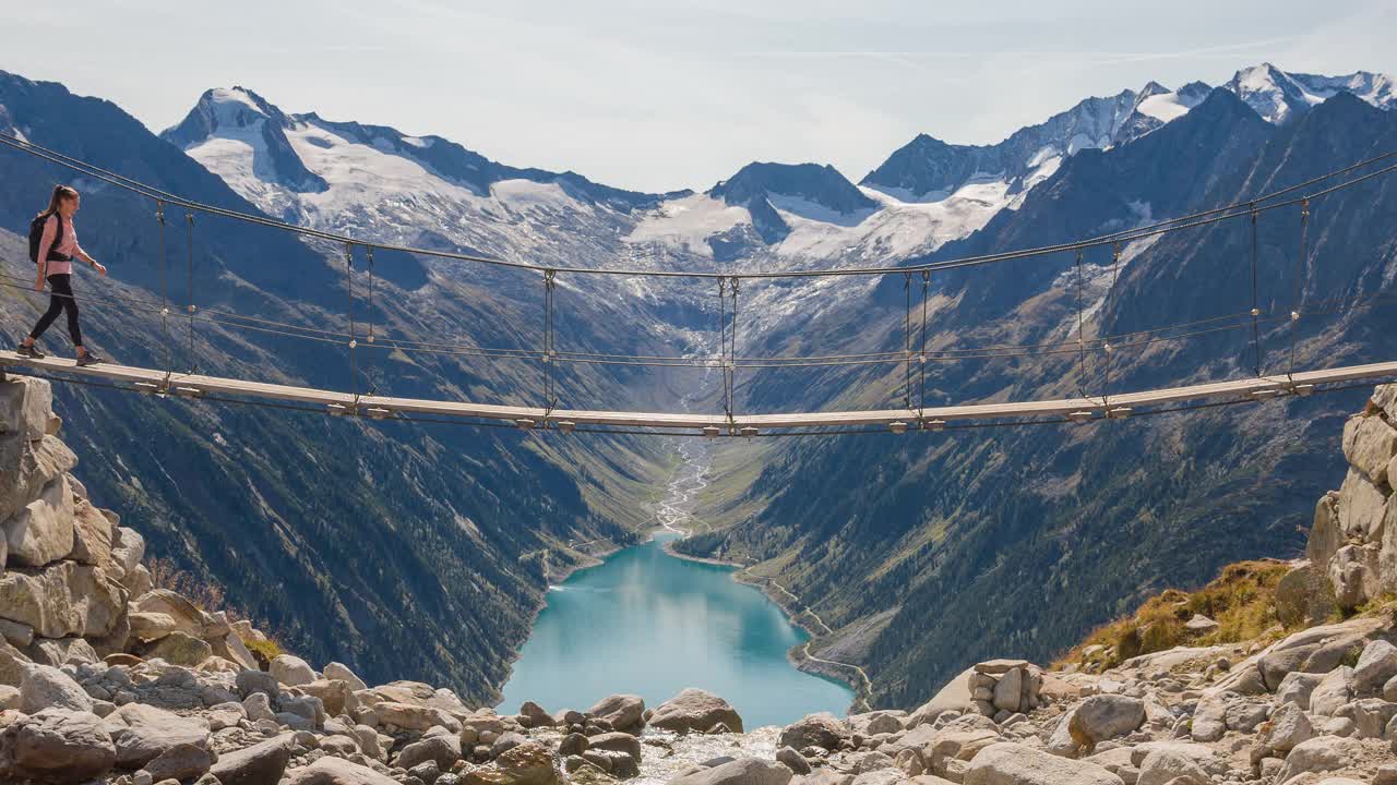 年轻的女徒步旅行者穿过淡水小溪上的吊桥，在高山上欣赏雄伟的山谷湖的景色视频下载