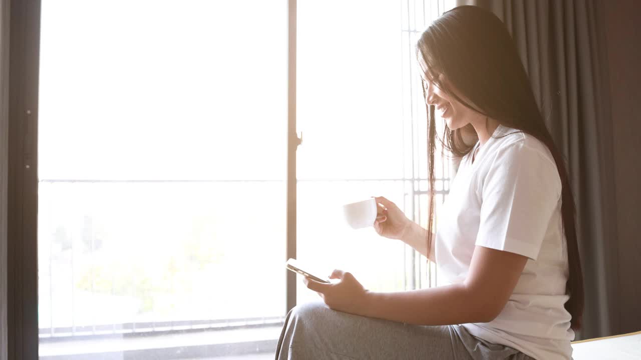 亚洲妇女坐在床上，喝一杯咖啡，同时使用手机在卧室在早上视频下载