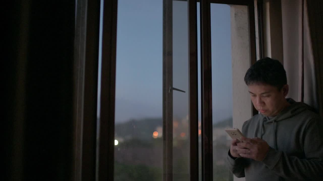 日落时分，亚洲华人中年男子穿着连帽衫在家中看智能手机，表情严肃视频下载