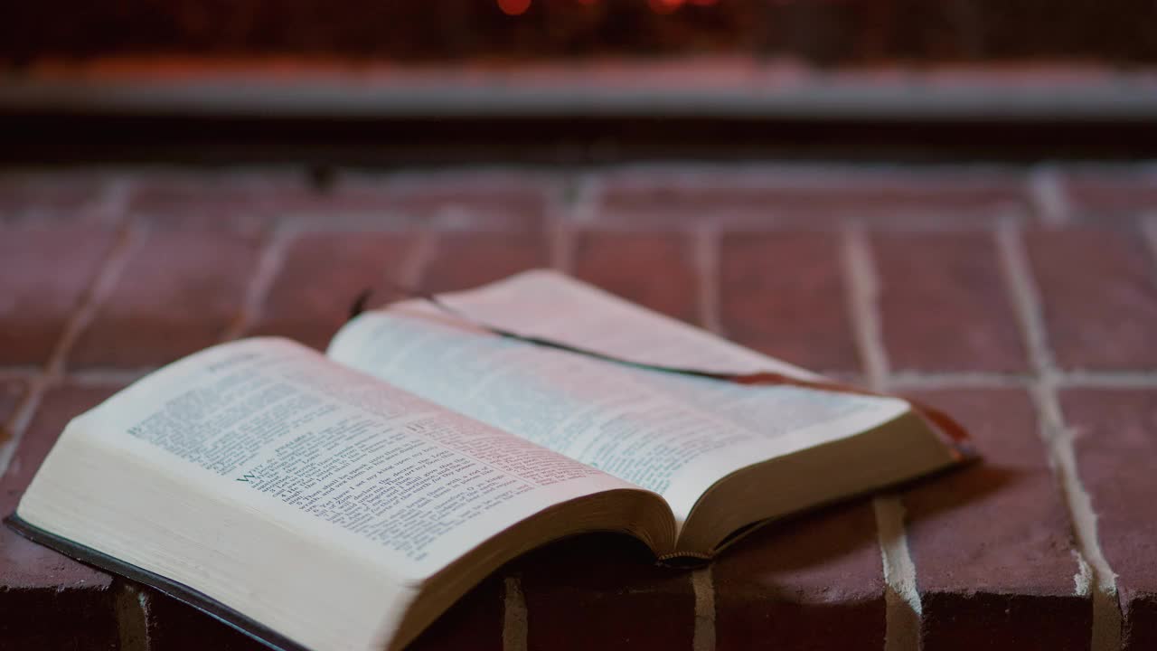 一本摊开在砖砌壁炉上的《圣经》，壁炉里的火焰投射出橘黄色的光芒，映照在《圣经》的书页上。视频素材