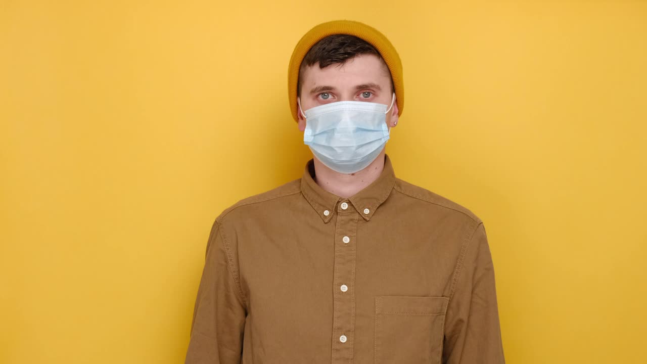 一名年轻的欧洲男子戴着医用防护口罩，穿着衬衫和无檐帽，孤立在黄色背景上，看着和展示大尺寸的广告版面视频素材