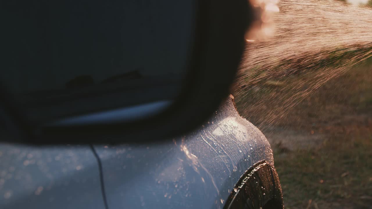 近距离观察水滴在汽车表面，而亚洲妇女清洗她的汽车用软管喷洒水在家里在日落时间。慢动作镜头。视频下载