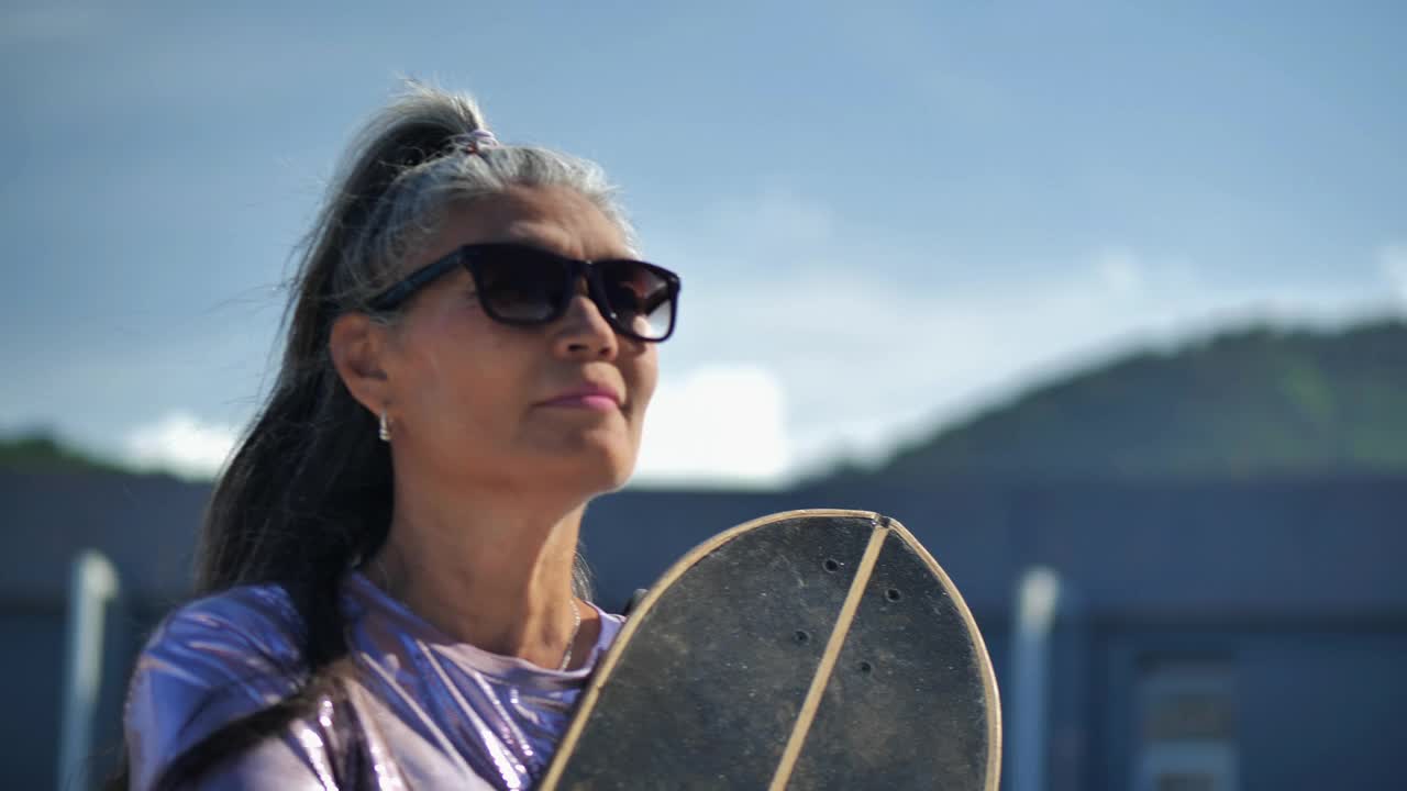 肖像拉丁美洲高级运动妇女戴太阳镜和灰色头发的年龄53岁持有滑板站在一个阳光明媚的日子。年龄与态度:高级肖像概念。视频下载