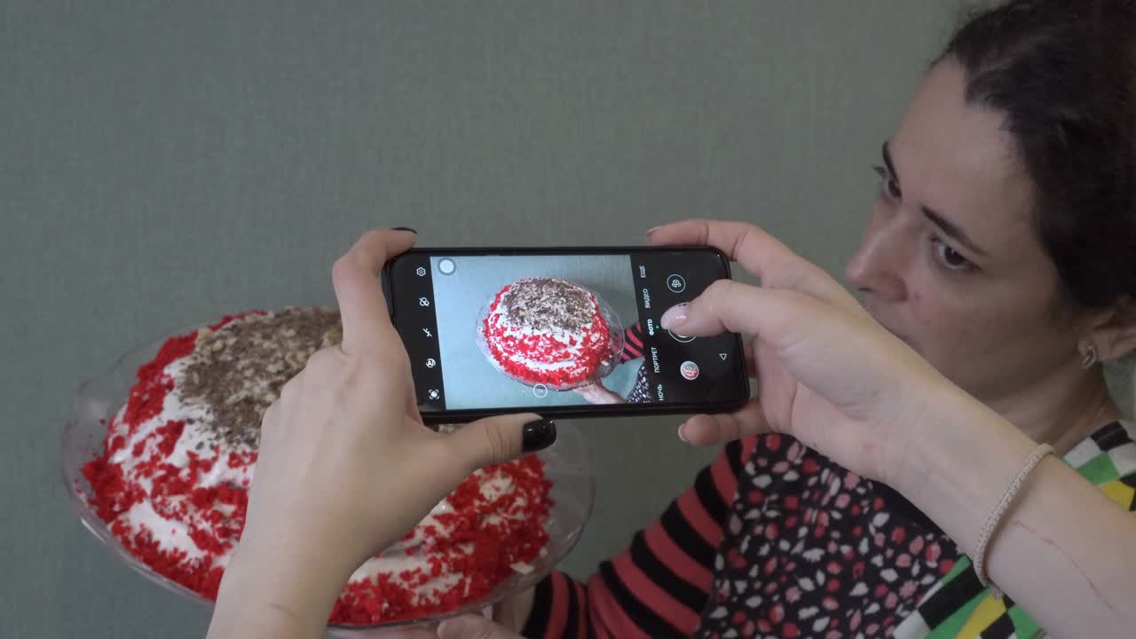 妈妈和女儿用智能手机拍食物的照片，一个漂亮的红色蛋糕在家里做。隔离期间居家经营的概念。特写镜头。从后面俯视。4 k。视频下载