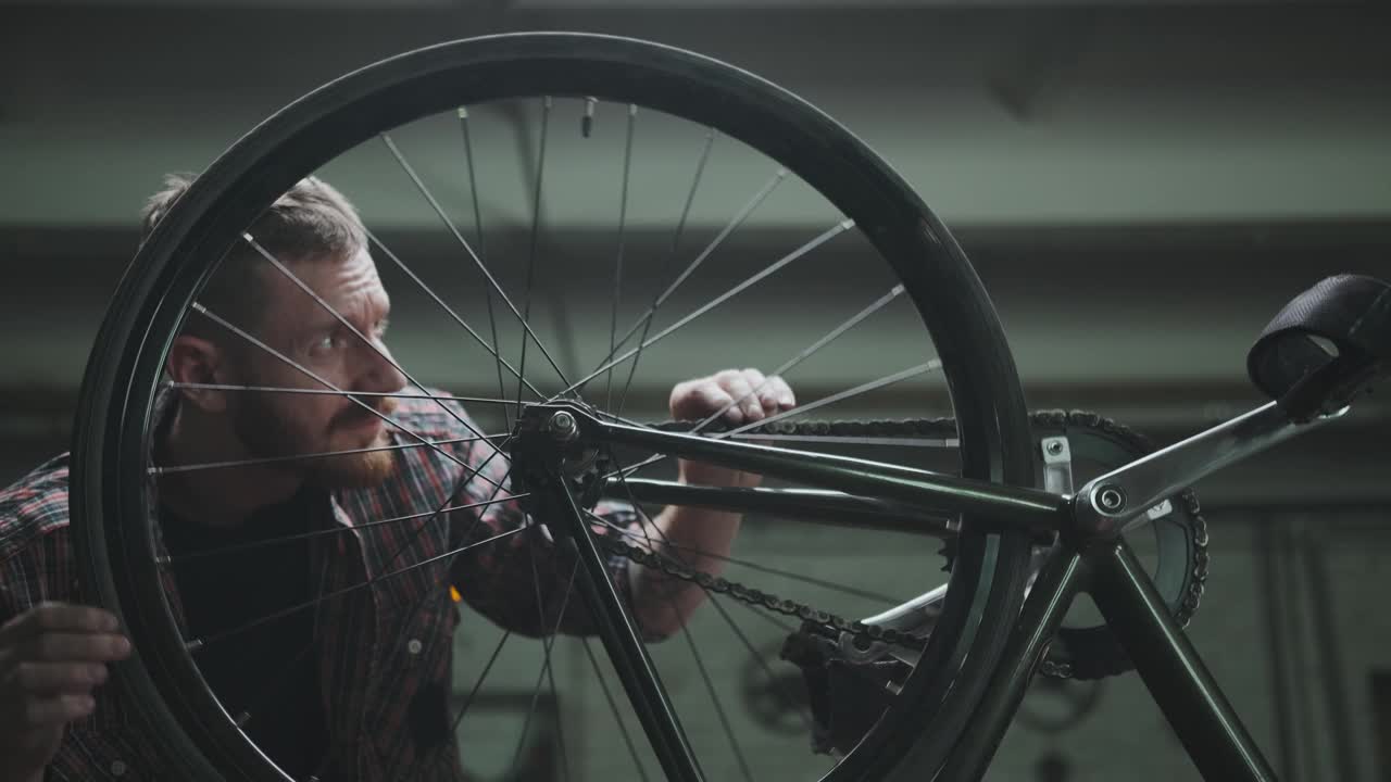 一名穿着格子衬衫的男性骑自行车者在车库里修理一辆自行车，转动轮子检查它。视频下载