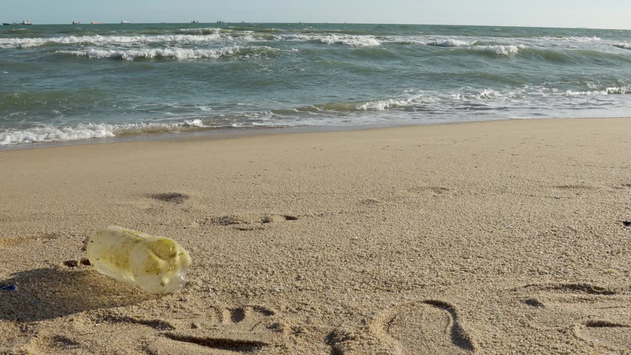 近距离观看4K视频，清晰的塑料瓶被丢弃在美丽的海滩和强大的海浪，看到地平线上的工业船只显示废物污染海洋的环境问题。视频下载