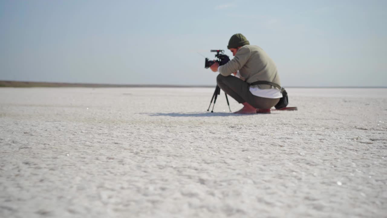 摄像师在工作中左右移动。一名男子坐在像沙漠一样的干燥矿湖的白色盐碱滩上用三脚架拍摄视频下载