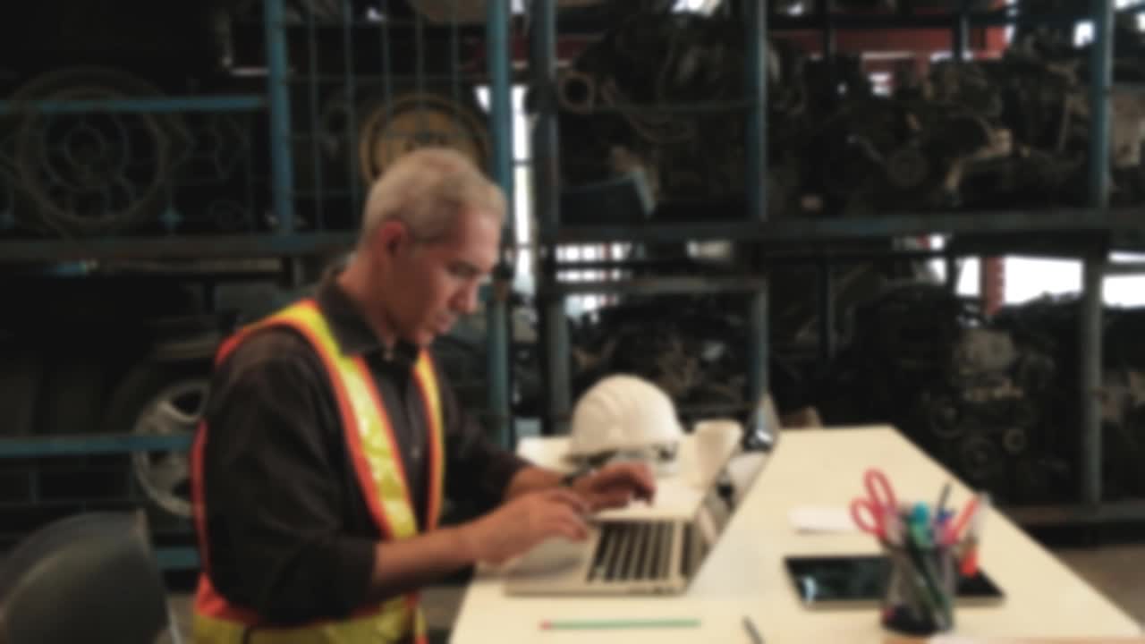 模糊焦点VDO:高加索男性工程师集中工作在桌子上。在备品仓库用笔记本电脑检查汽车发动机和机械的订单和库存。视频下载