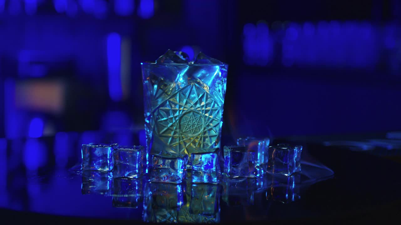 酒保点燃酒精鸡尾酒，蓝色背景。鸡尾酒杯中的火焰在慢镜头中燃烧，酒精饮料中的冰块，酒保制作饮料。视频素材
