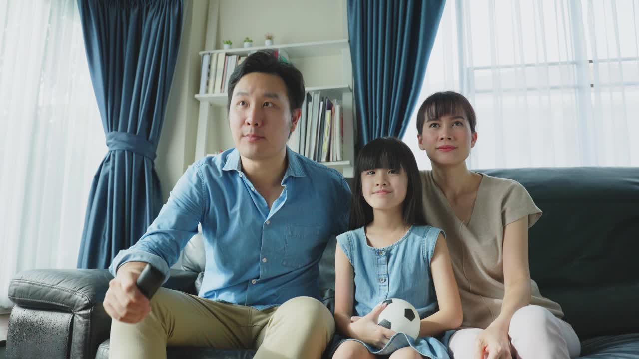 亚洲幸福的父母和女儿坐在家里客厅的沙发上。兴奋的家庭，年轻的小女孩球迷与父母拿着足球看电视体育比赛，庆祝球队的胜利。视频购买