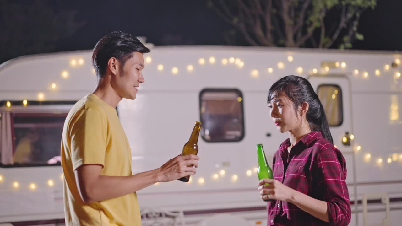 亚洲年轻男女旅行者朋友正在举行户外节日聚会。夫妻俩一起喝一瓶啤酒，晚上一起开怀大笑。旅行自然，露营车露营。视频素材