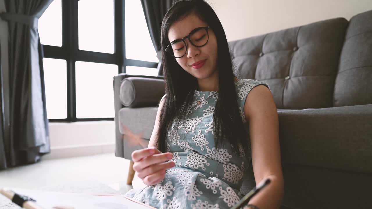 美丽的年轻亚洲女艺术家坐在地板上和素描与画板在客厅视频下载