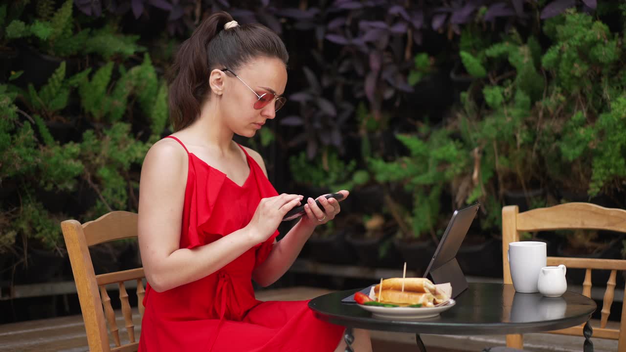 侧视图的专注年轻的女人在太阳眼镜和红色连衣裙工作在线坐在咖啡馆在美丽的地中海国家。专注于平板电脑和智能手机上的自由职业者信息传递。视频素材