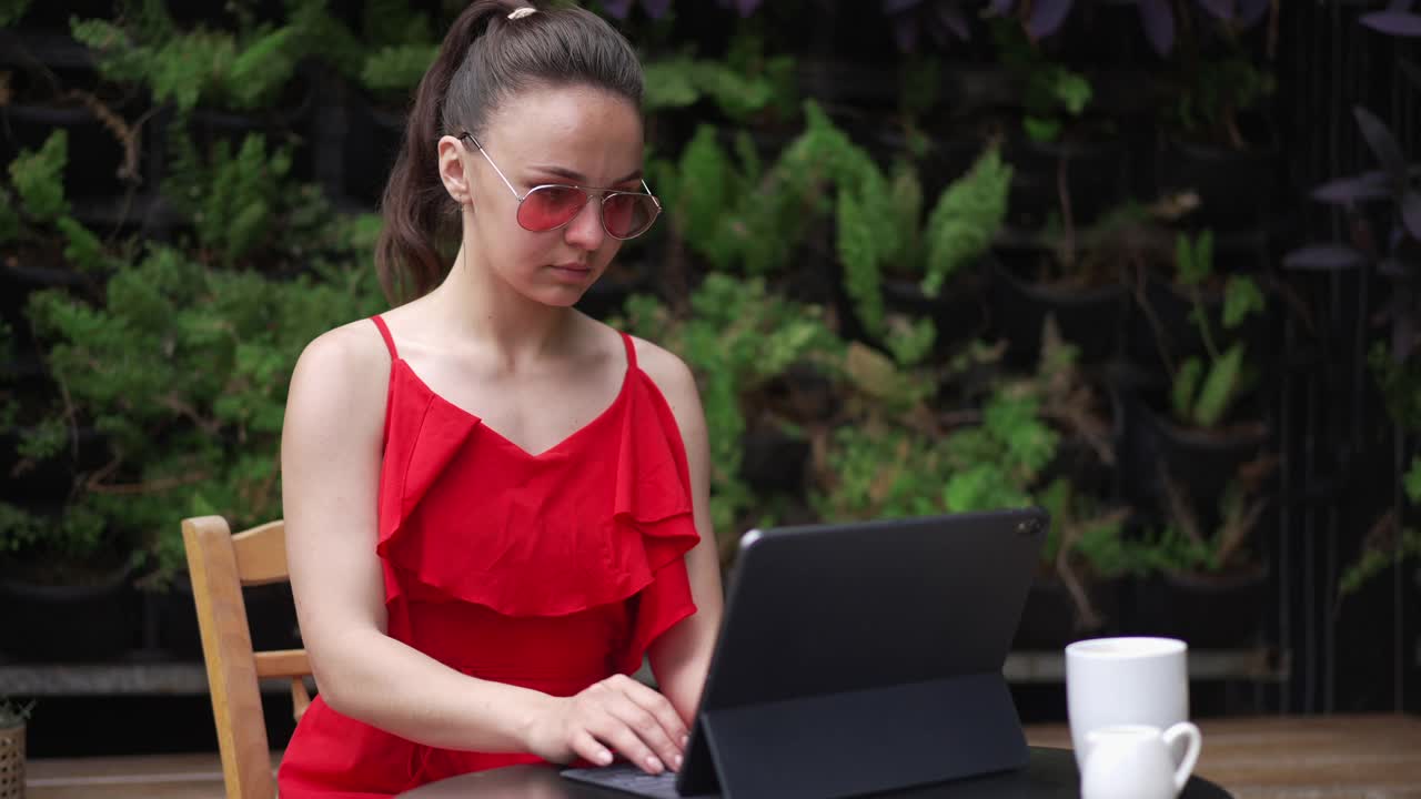 一名年轻女子在平板电脑上查看电子邮件，而她的身份是在户外咖啡馆送食物的陌生服务员。正面的白种人自由职业者的肖像工作消息，在线冲浪互联网。方便的概念。视频素材