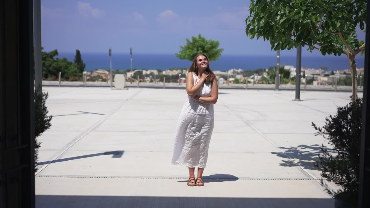 宽镜头的照片，兴奋的高加索年轻美丽的女人站在阳光下地中海旅游胜地微笑欣赏自然。快乐的白衣女士在户外度假。视频素材