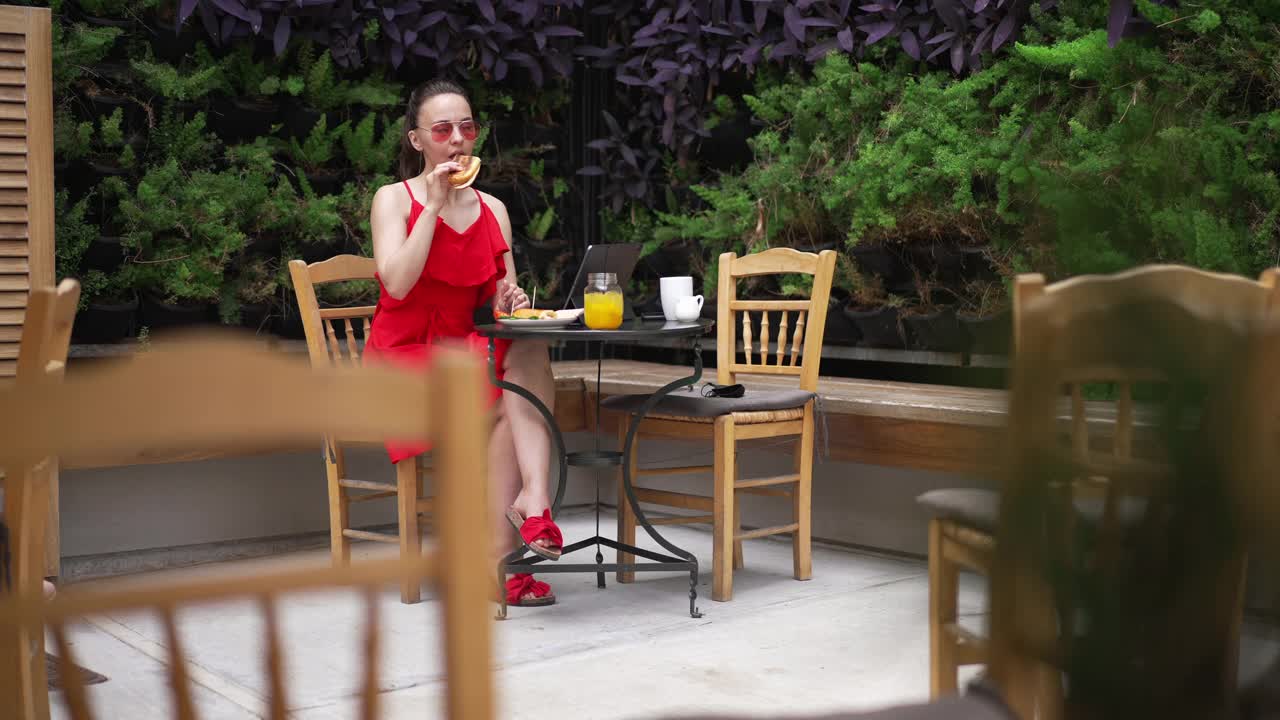 广角拍摄的快乐美丽的年轻白人女子穿着红裙子和太阳镜吃汉堡，喝鸡尾酒坐在咖啡馆的户外。自信优雅的自由职业者与平板电脑度假的肖像。视频素材