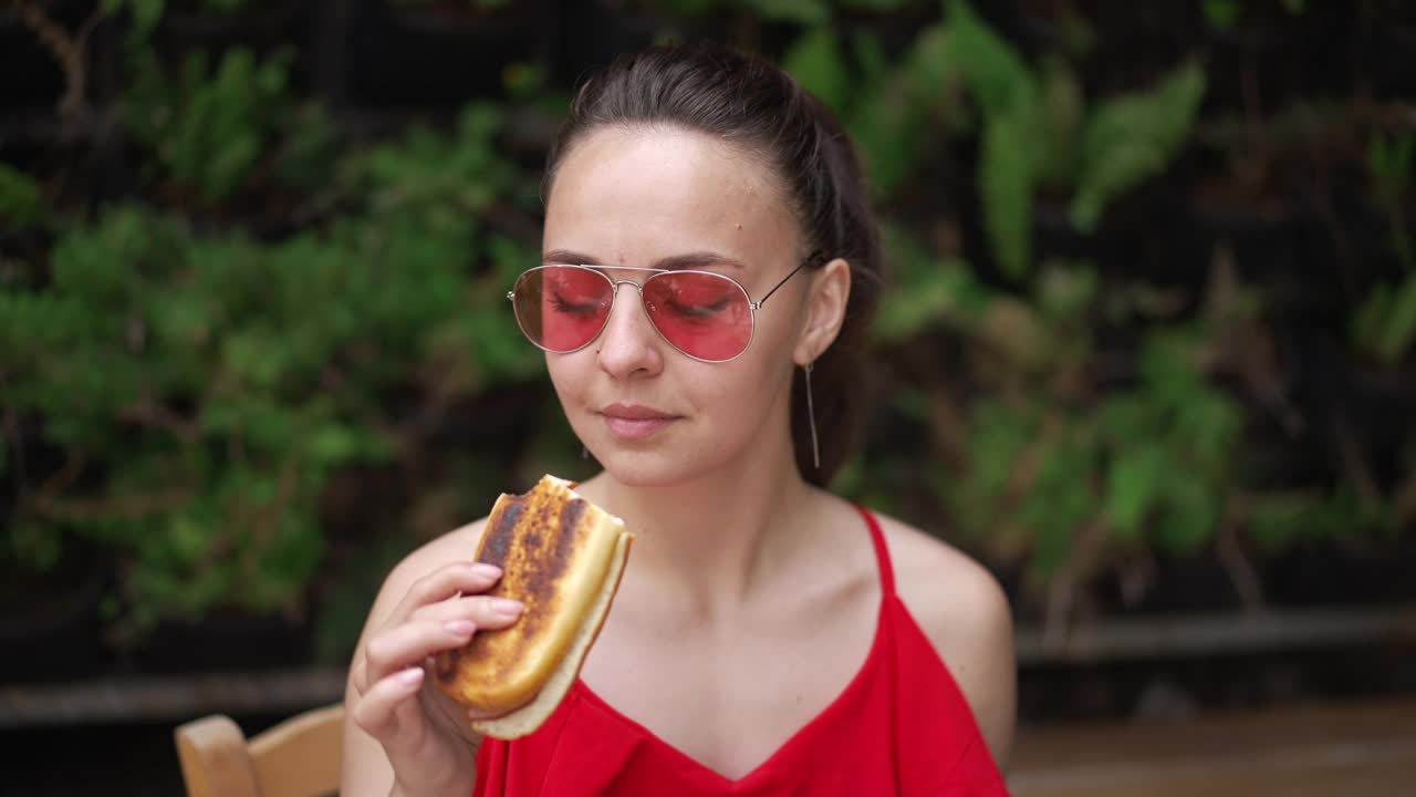 年轻的女游客肖像享受美味的汉堡在户外咖啡馆度假。美丽苗条自信的白人女性休息在度假村的户外。缓慢的运动。视频素材