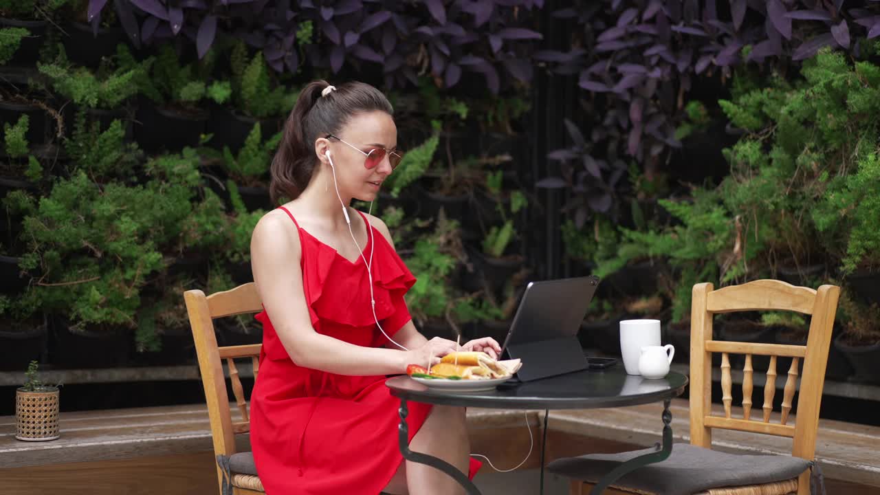 微笑，开朗，美丽苗条的白人年轻女子戴着耳机，随着音乐跳舞，坐在咖啡馆里用平板电脑聊天。积极自信的自由职业者在假期在线工作。视频素材