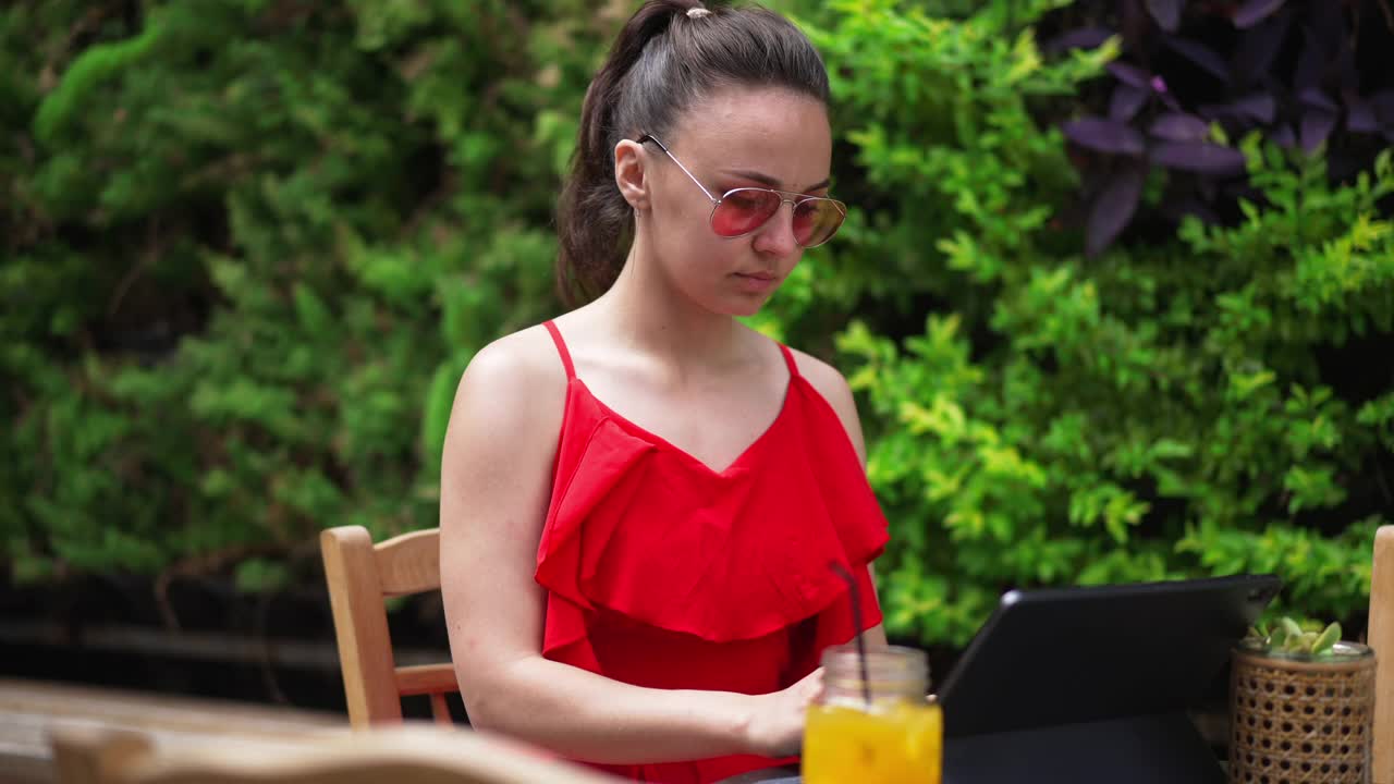 照片中的年轻美丽的女人戴着太阳镜，坐在户外的夏日咖啡馆里，用平板电脑在网上聊天。集中分析电子市场思维的白人女性自由职业者。视频素材