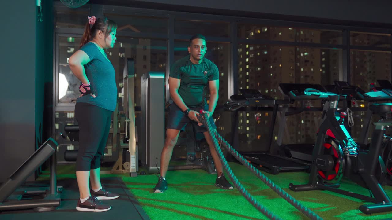 身体积极的亚洲华人中年女性观看她的私人教练展示正确的方法在健身房练习战斗绳视频素材