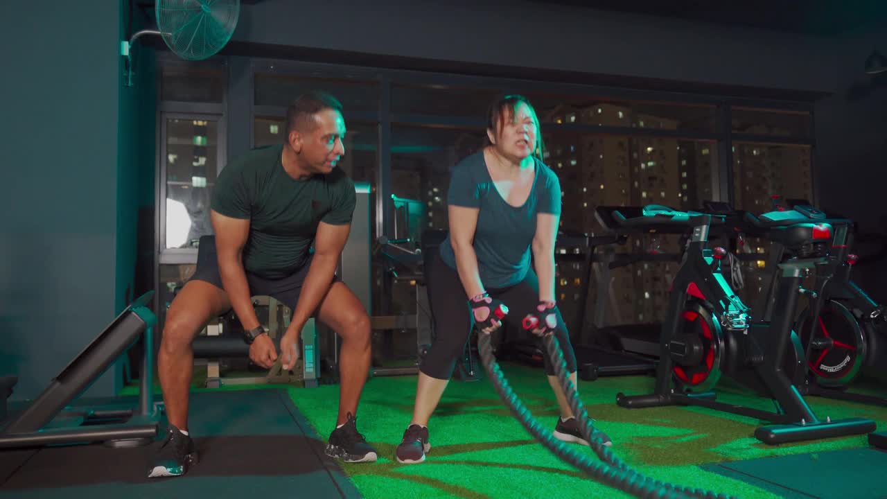 身体积极的亚洲华人中成年妇女在健身房练习战斗绳与她的印度男性私人教练视频素材