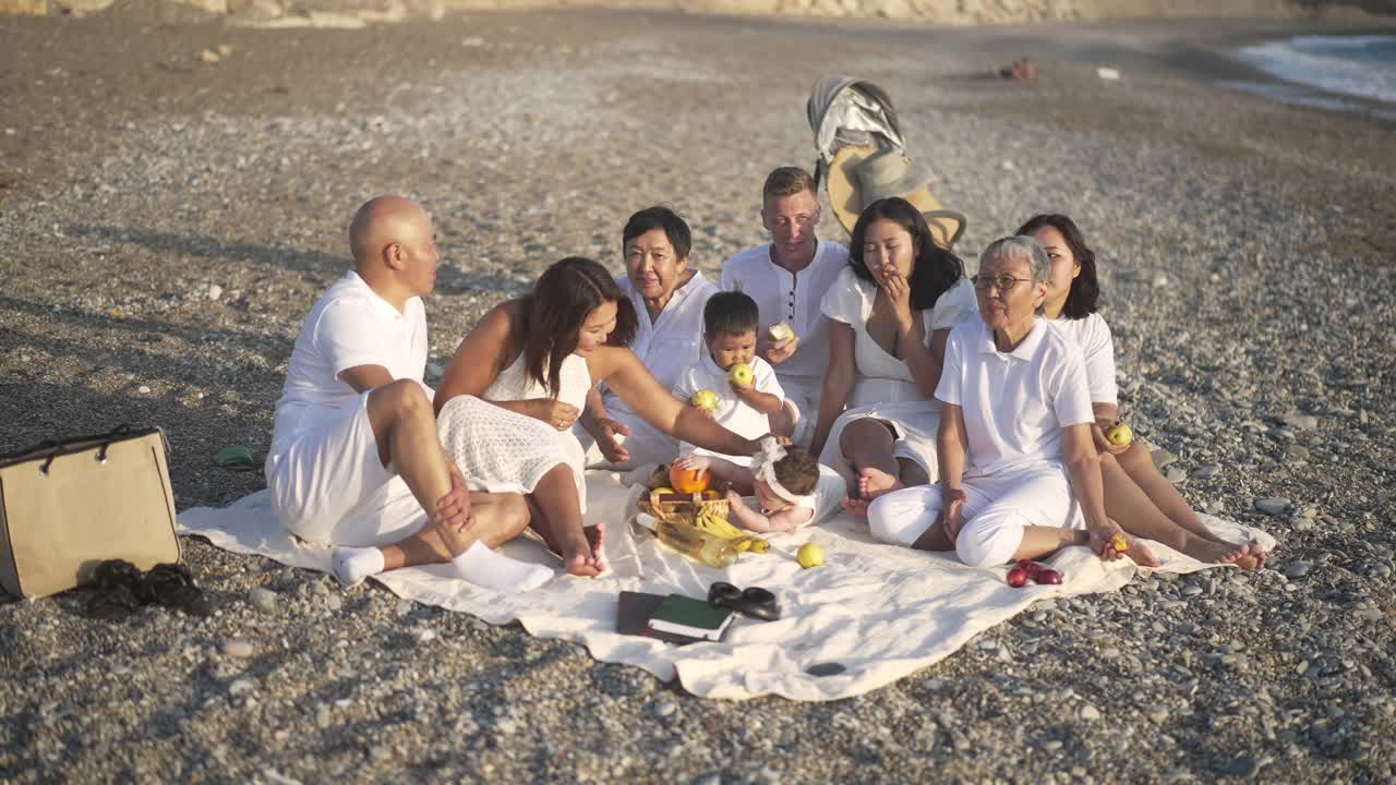 广角拍摄的快乐无忧无虑的亚洲家庭与白种人男子坐在毯子上岩石海岸聊天笑慢动作。放松的男人，女人和孩子享受家庭度假胜地。视频素材